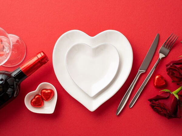 Foto von herzförmigen Tellern, Besteck, Weinflaschen, Rosengläsern und Schokoladenpralinen auf isoliertem rotem Hintergrund
